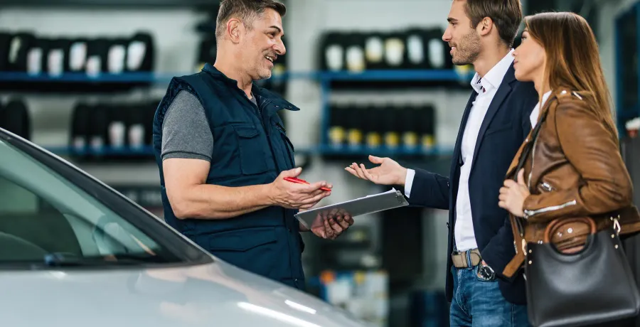 Mecânico de autocenter conversando com um casal de clientes ao lado de um carro, com prateleiras de pneus ao fundo