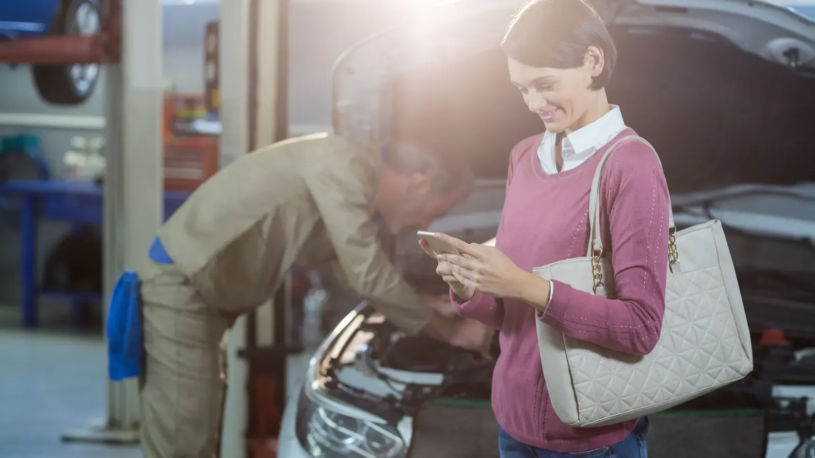 Cliente usando o celular dentro de uma oficina enquanto o mecânico trabalha no carro